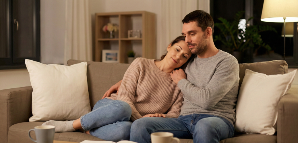 זוג יושב מחובק ורגוע על הספה בסלון מואר וחמים, הממחיש בניית אינטימיות רגשית, תחושת ביטחון ומגע מקרב ללא לחץ מיני כחלק מתהליך של גישור על פערים בזוגיות.
